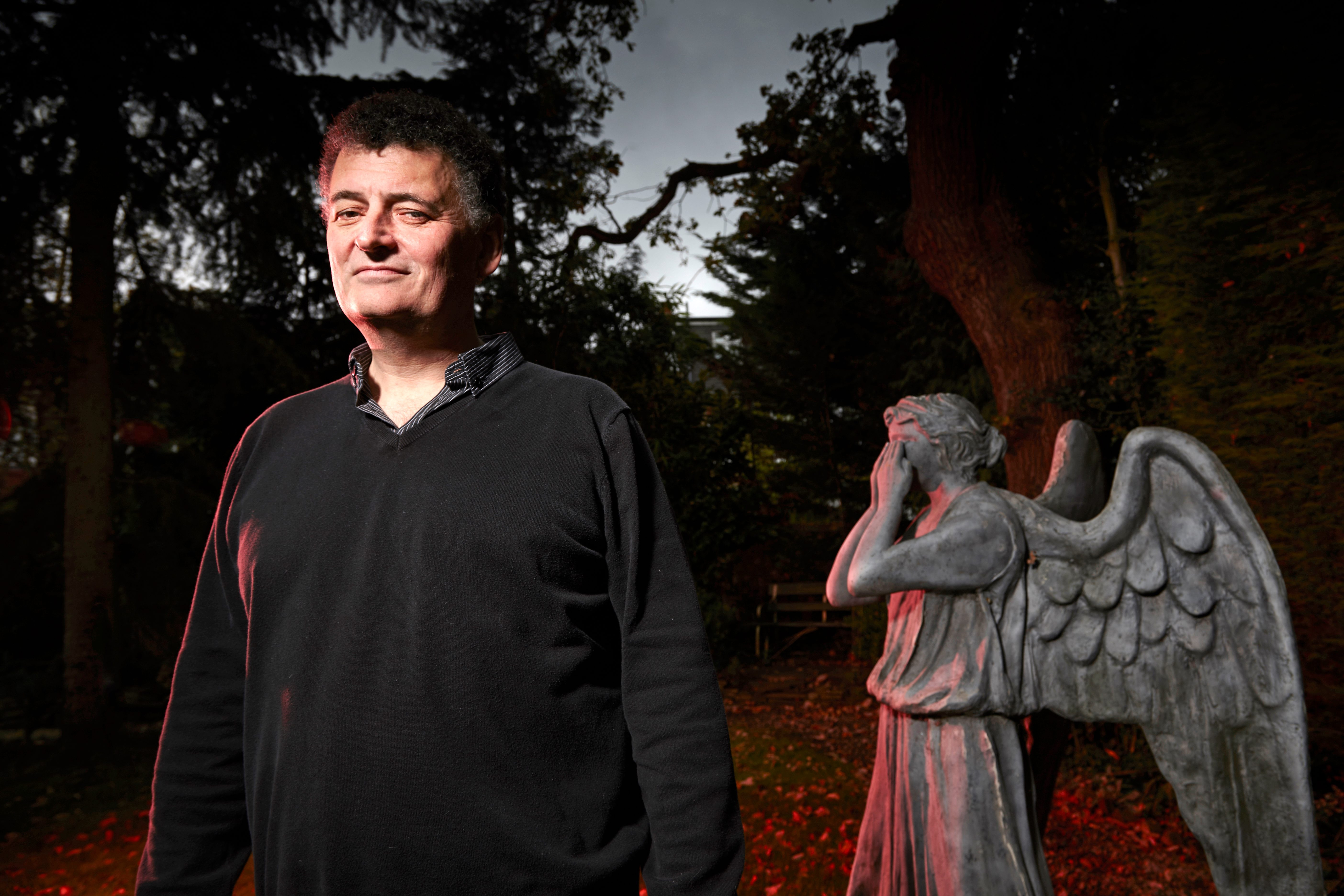 Steven Moffat standing wearing a grey jumper, with a Weeping Angel in the background
