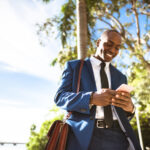 A smiling businessman pauses in his on-foot commute to respond to a message on his cell phone.