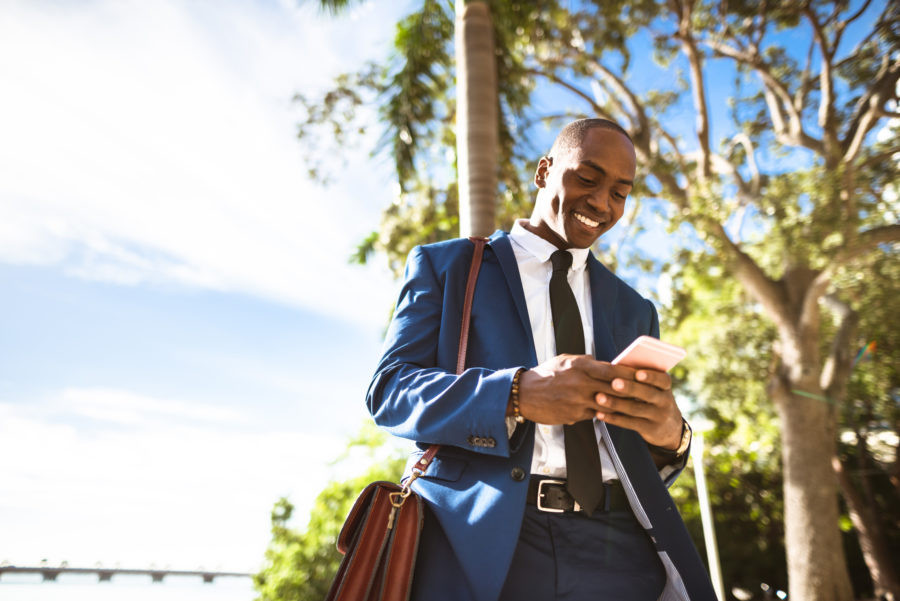 A smiling businessman pauses in his on-foot commute to respond to a message on his cell phone.